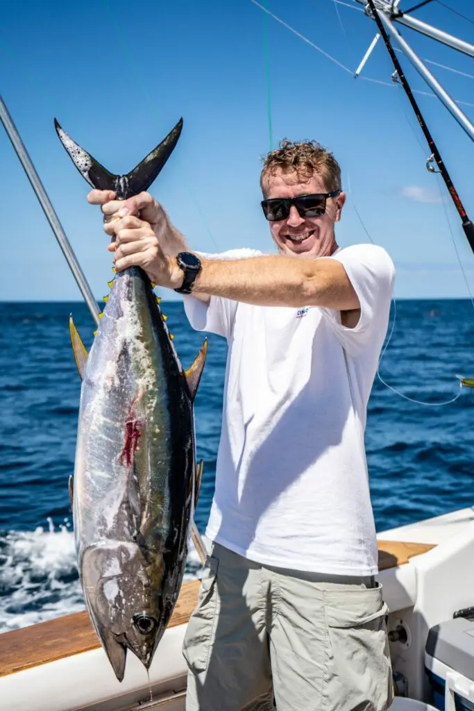 A smiling sportfishing guest at Chino Salsa stands on a boat holding up a large yellowfin tuna against a backdrop of bright blue ocean and sky.