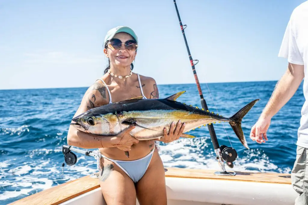 Chino Salsa's guest on the sportfishing boat smiles while holding a large yellowfin tuna, with bright ocean waves and a clear blue sky in the background.
