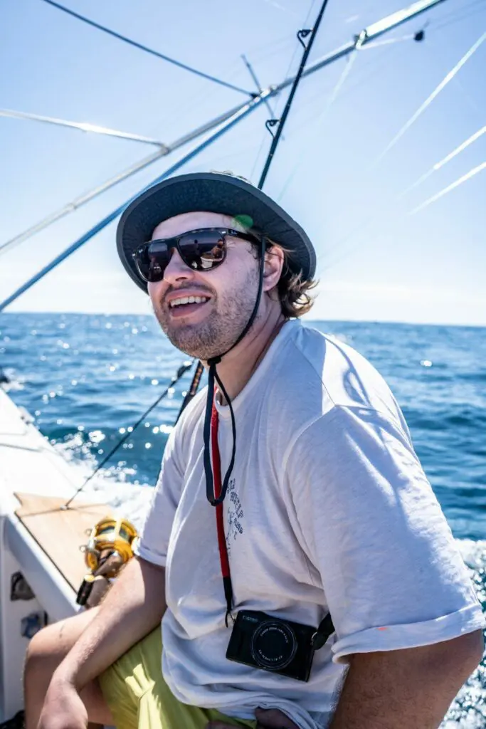 A smiling guest sits on the edge of chino salsa sportfishing boat in Flamingo waters, wearing sunglasses and a sun hat, with bright blue ocean and clear sky behind them.