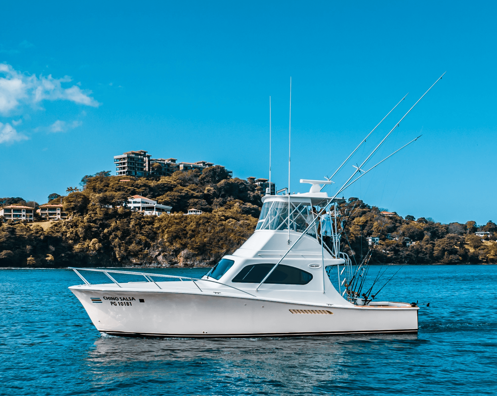 Chino Salsa sportfishing boat floating on calm blue water with lush hillside homes rising behind it under a clear, bright sky in Costa Rica